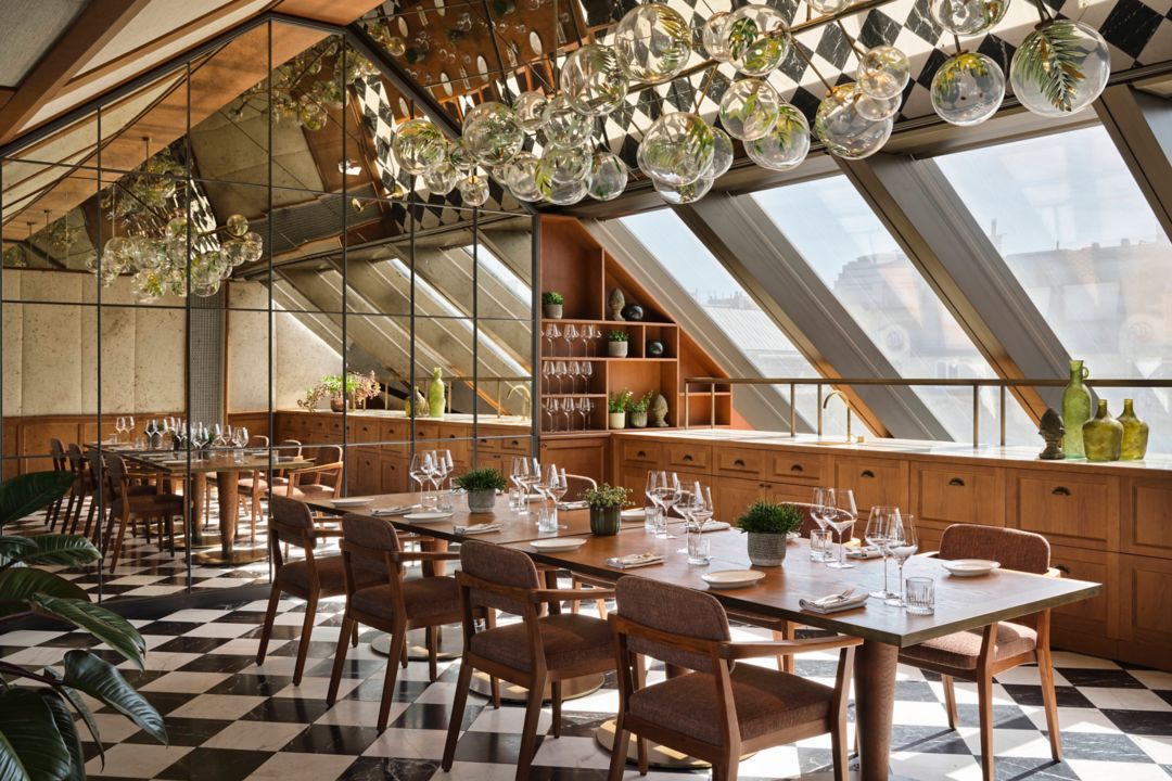 A dining table under hanging glass lights and  large slanted windows.